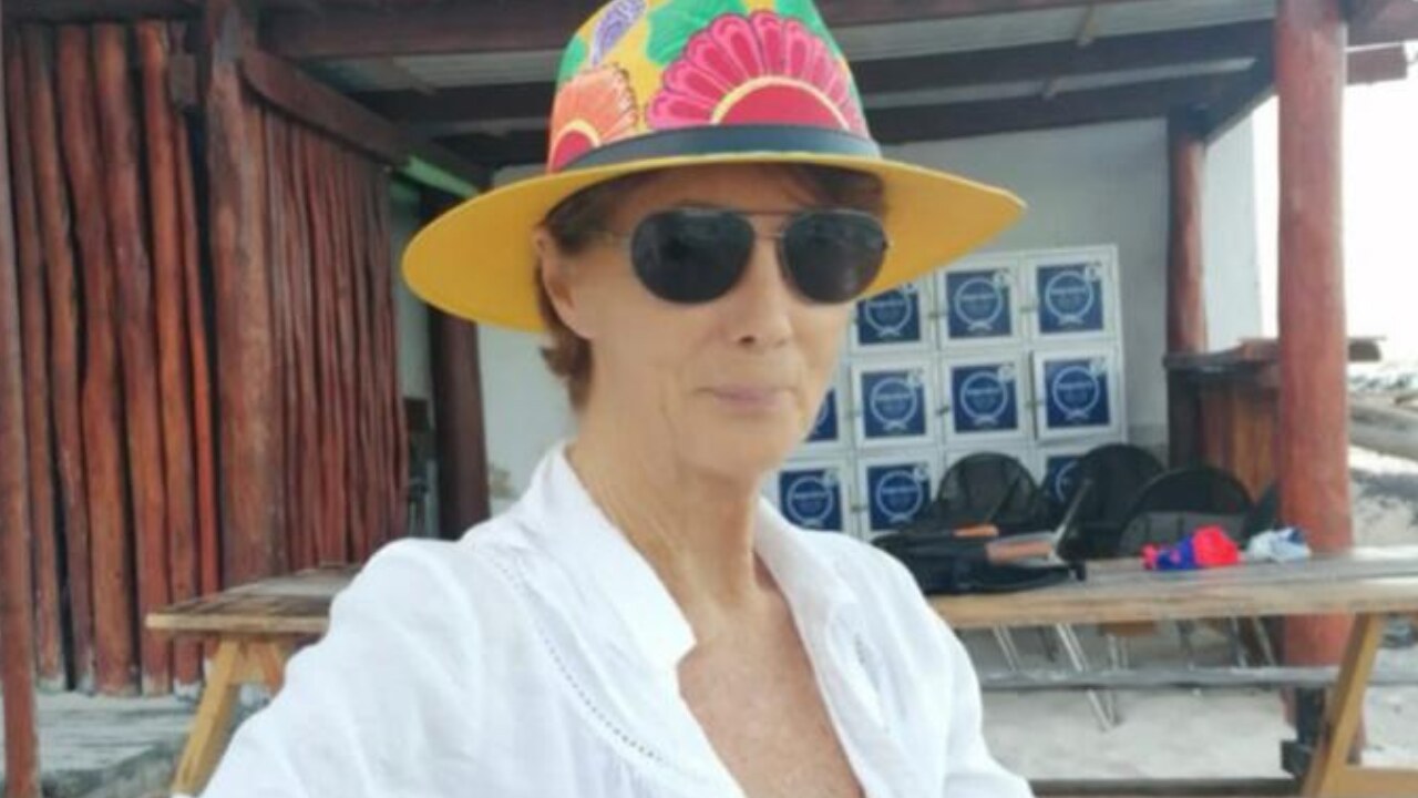 Barbara Crawley wearing a bright sun hat and sunglasses, seated outdoors
