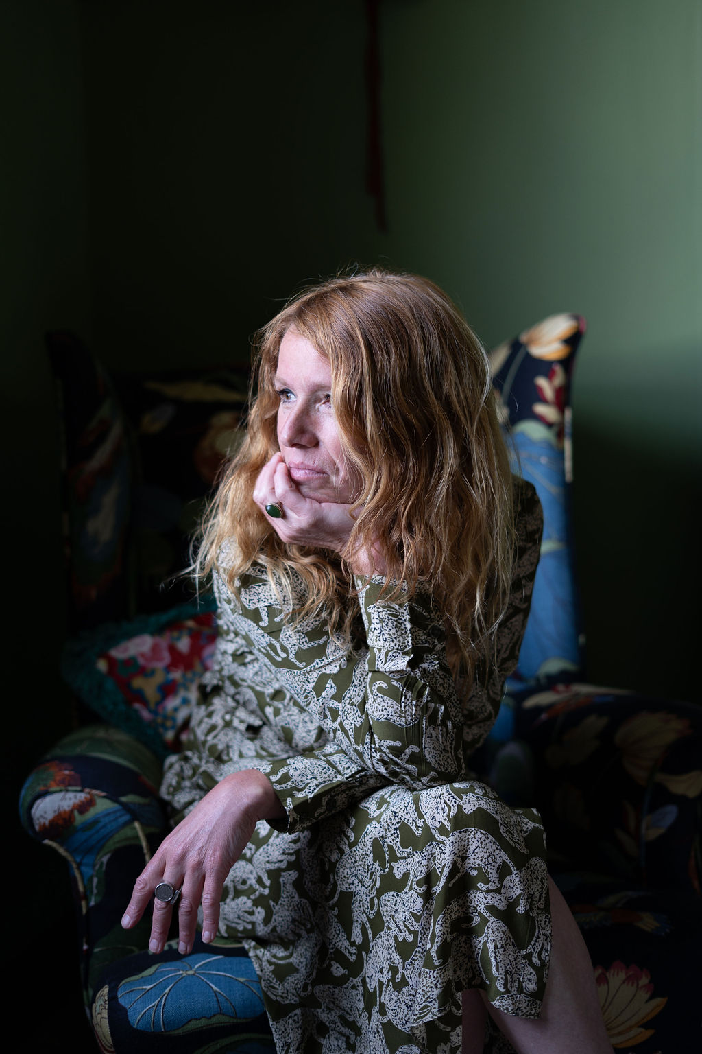A woman with wavy auburn hair seated thoughtfully in an armchair, her hand resting against her cheek