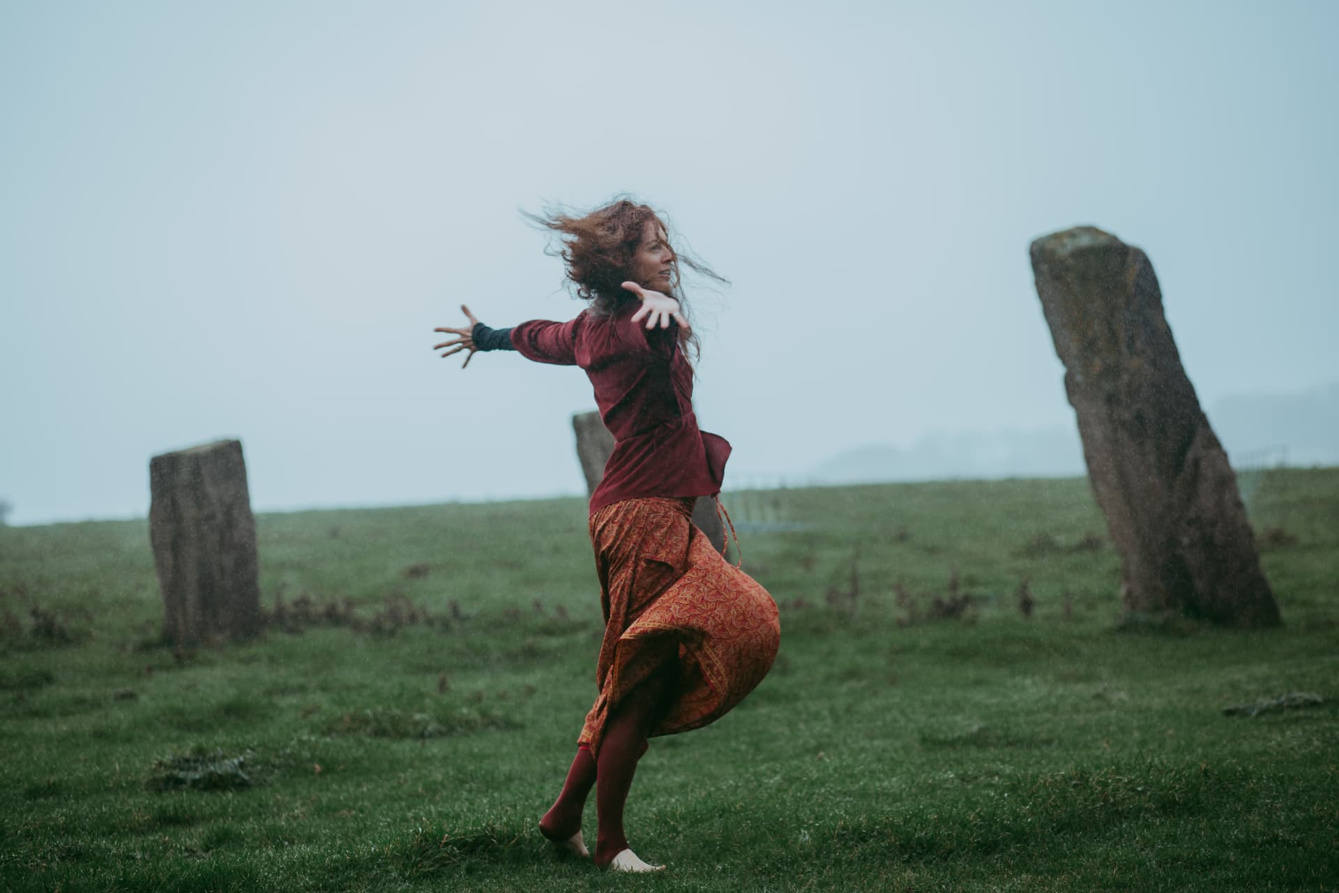 A woman with her arms open, dancing among ancient standing stones in an open landscape