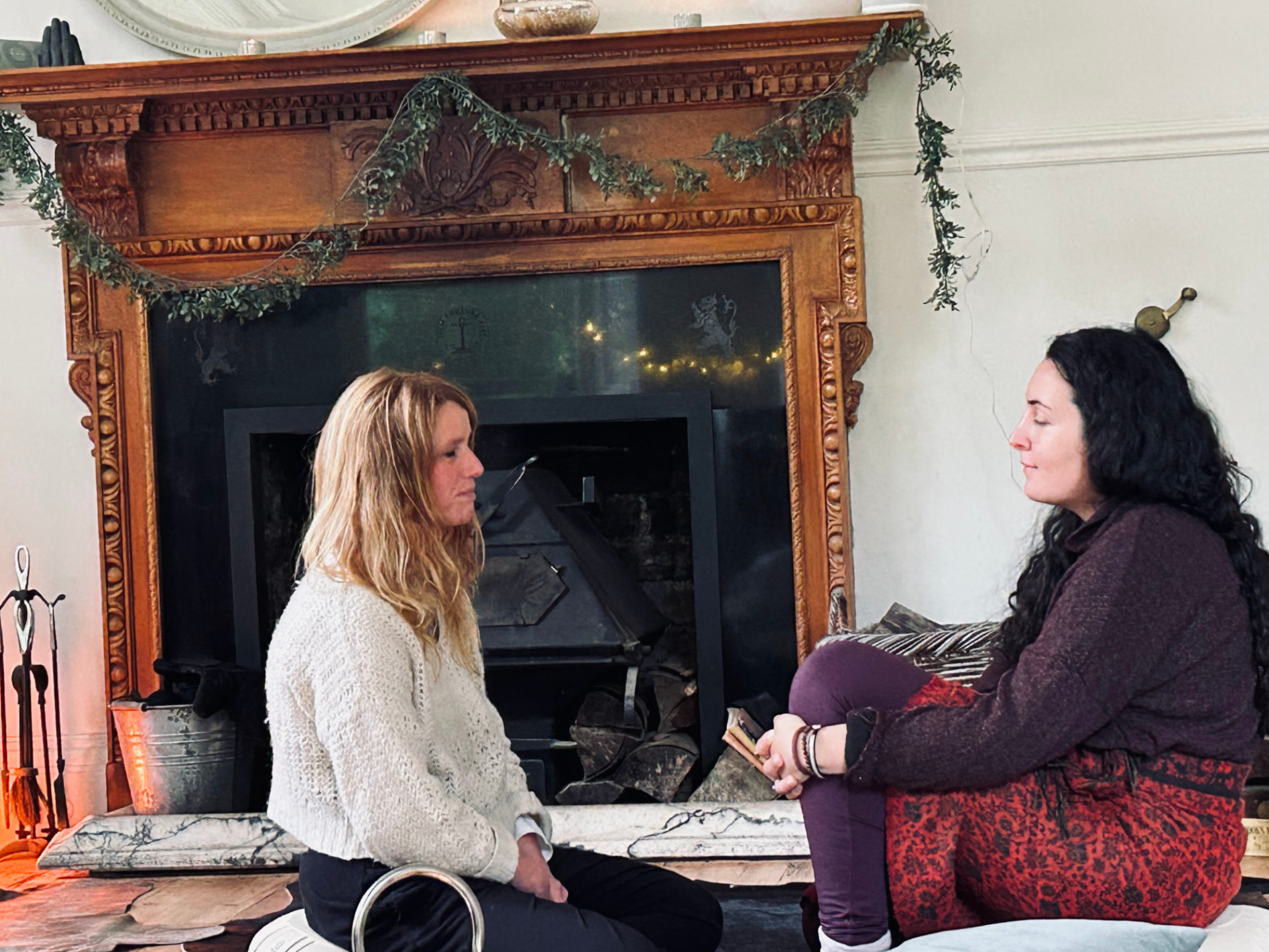 Two women sitting on cushions facing one another in front of a carved wooden fireplace draped with a green garland