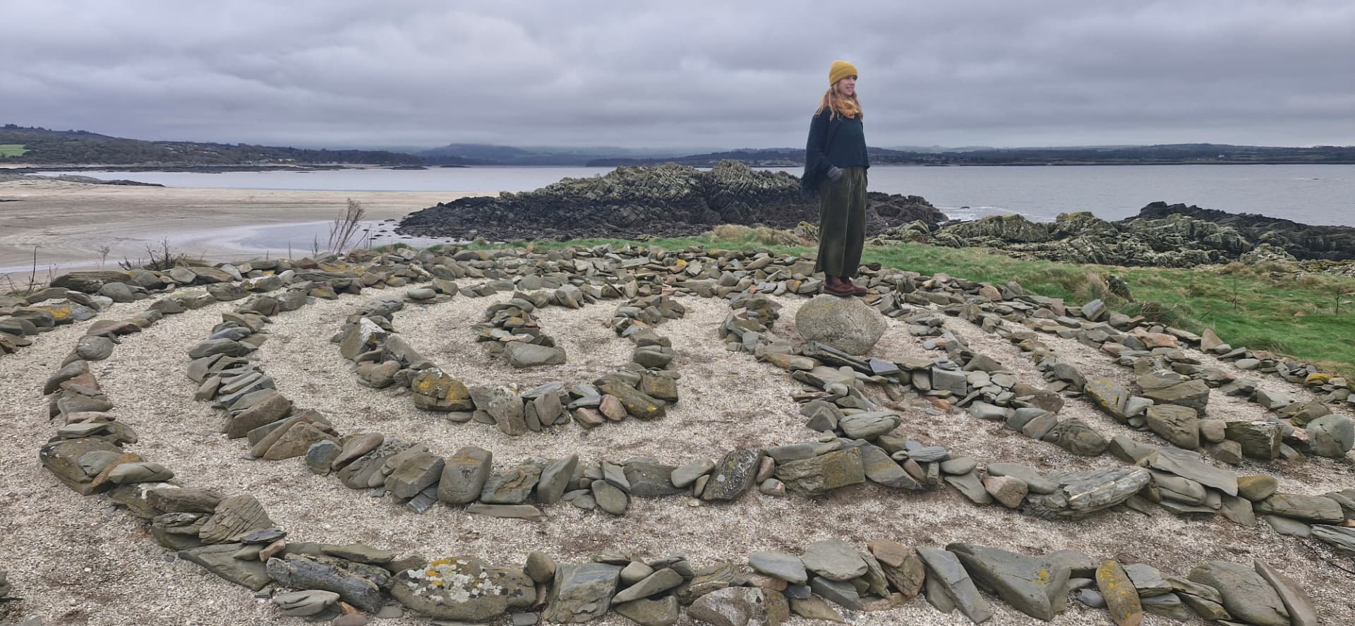 A woman standing on the centre stone of a coastal stone spiral, looking out over a bay
