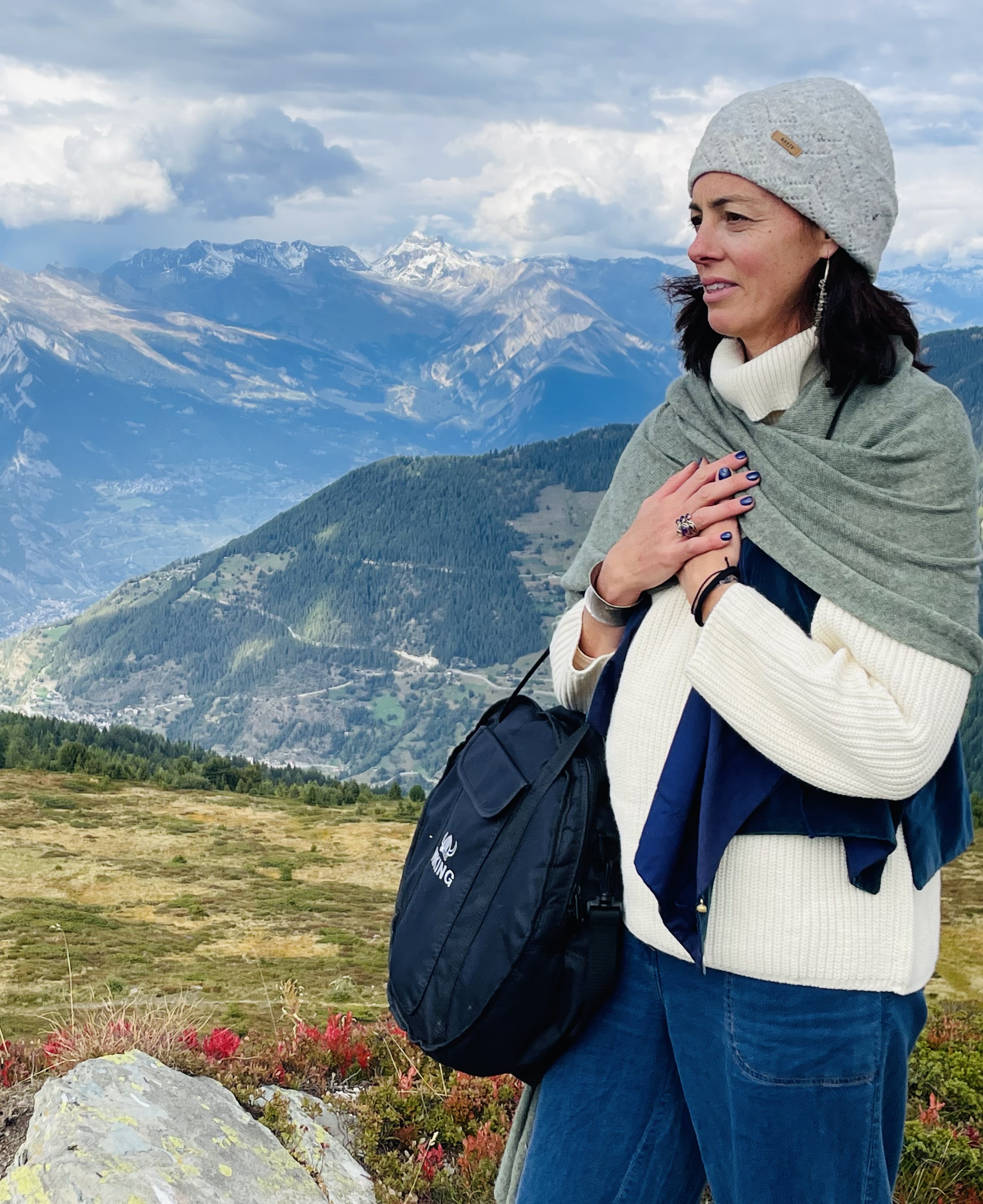 A woman in a cream knit turtleneck and sage shawl standing on a hillside, her hands resting on her chest, a wide mountain valley spreading behind her